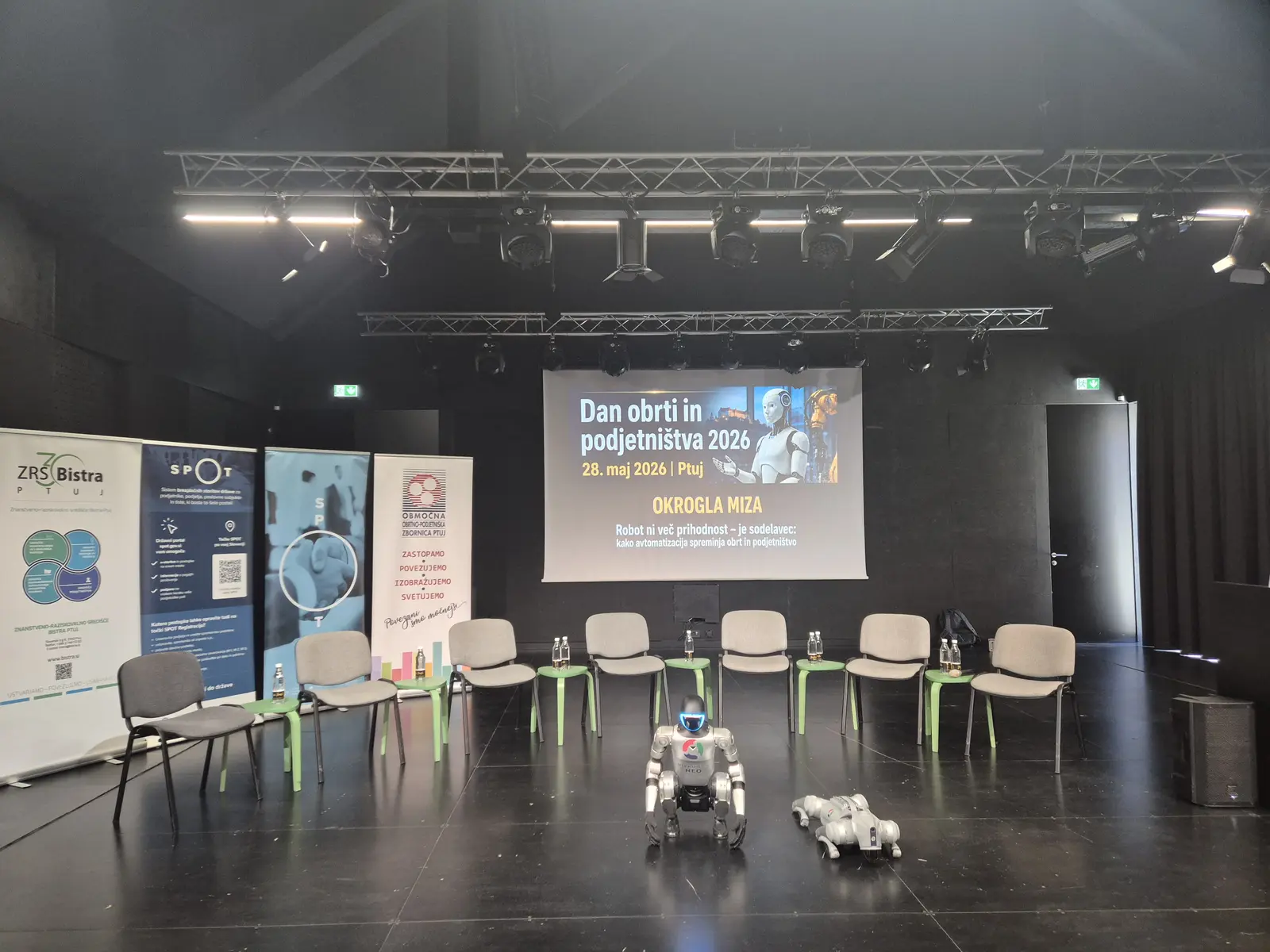 Round-table venue: chairs in a semicircle, the »Dan obrti in podjetništva 2026« projection, and a humanoid and quadruped robot.